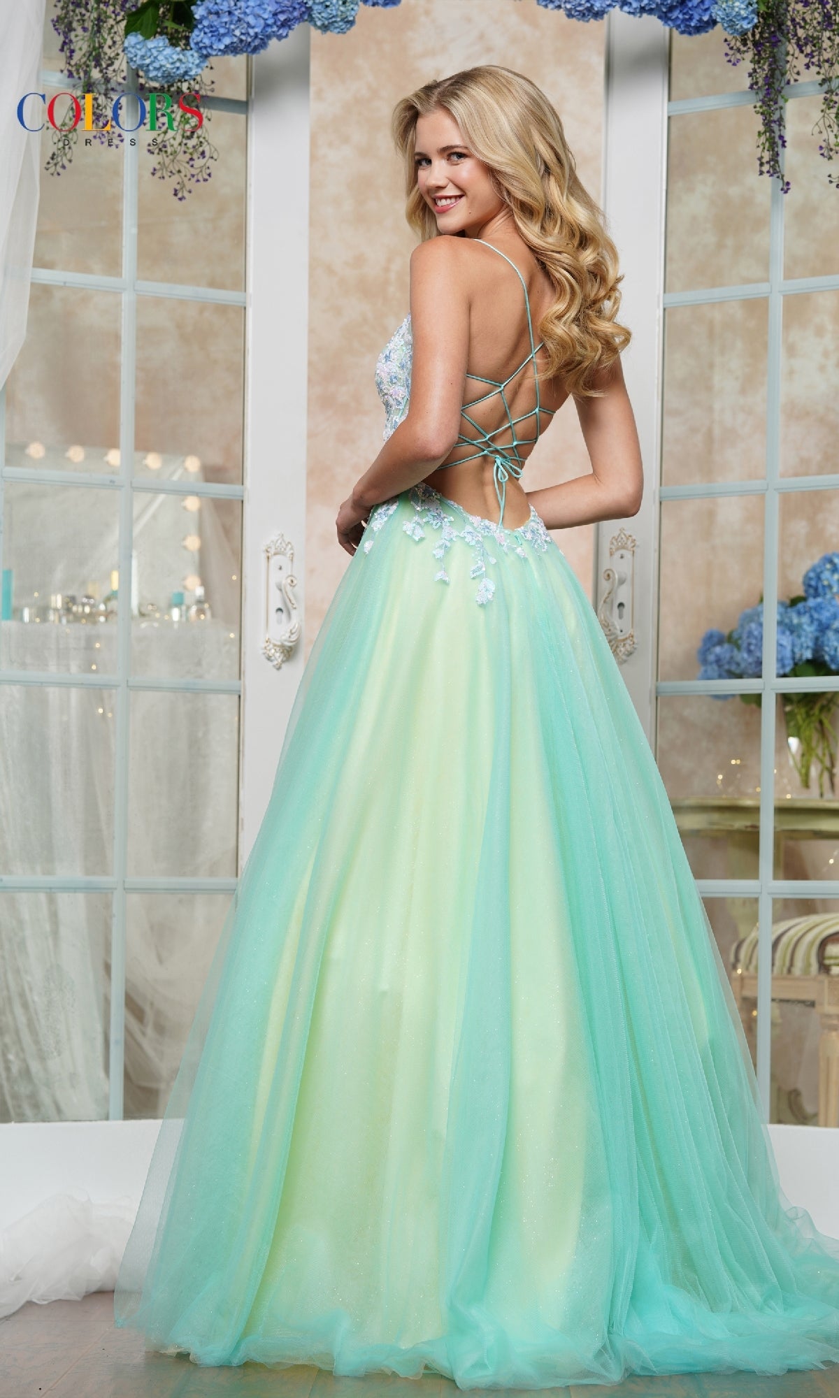A woman smiles in a decorated room wearing the Strappy-Back Prom Ball Gown: Colors Dress 3497, a pastel green and blue evening gown with an open lace-up back, surrounded by flowers and large windows.